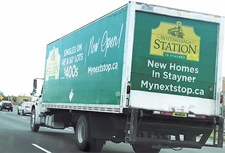Mobile billboard truck driving on highway with green and yellow ads on it side and back for MacPherson Home Builders.