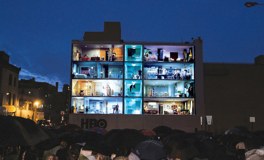 An image of an HBO outdoor advertisement that is a projection of the inside of an apartment building. The fake apartments are lit up to display what's going on inside each.
