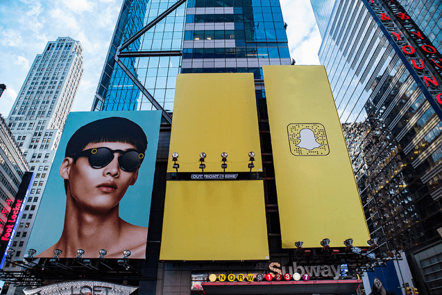 An image of an ad for snapchat. Three of the big billboards are yellow, one has a QR code of the snapchat app and one billboard has a model wearing Spectacles.