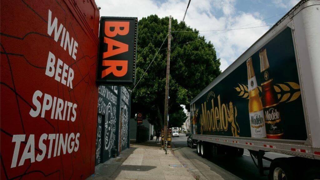 An image of a truck with an ad on the side of it for Modelo beer. The truck is parked in front of a bar that has "wine, beer, spirits, tastings" in bold letters on the side of it. 