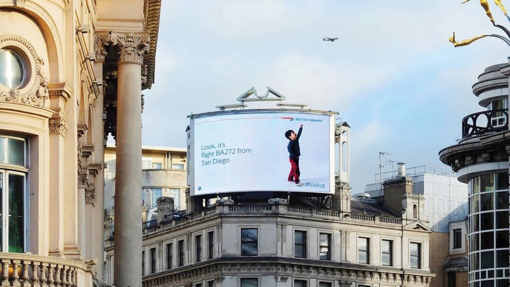 An image of a DOOH billboard ad for British Airways displaying a kid pointing up at the plane in the sky.