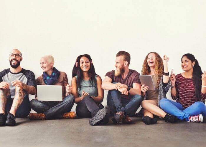 Group of people in their 30s sitting against a wall in a line. Laughing, talking and using electronics. 