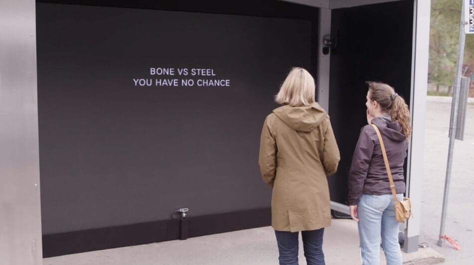 Two women stand in front of the digital billboard that reads, "bone vs. steel you have no chance."