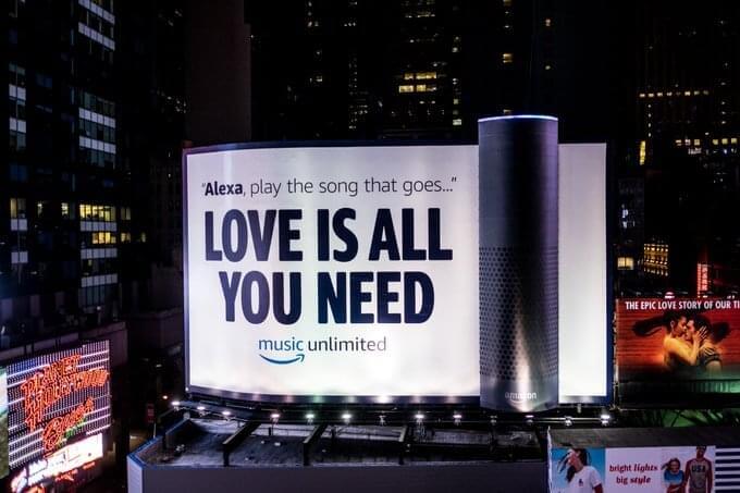 An image of a giant billboard for Amazon music that displays a gigantic speaker and says "Alexa, play the song that goes... Love Is All You Need".