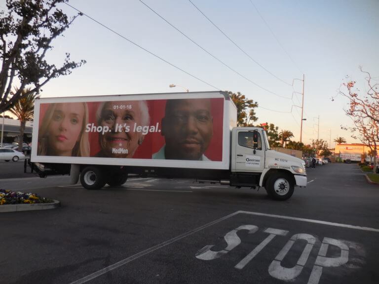 An image of a truck-side ad for MedMen. Has three faces on it, one a young girl, one an older woman smiling, and a middle-aged man.