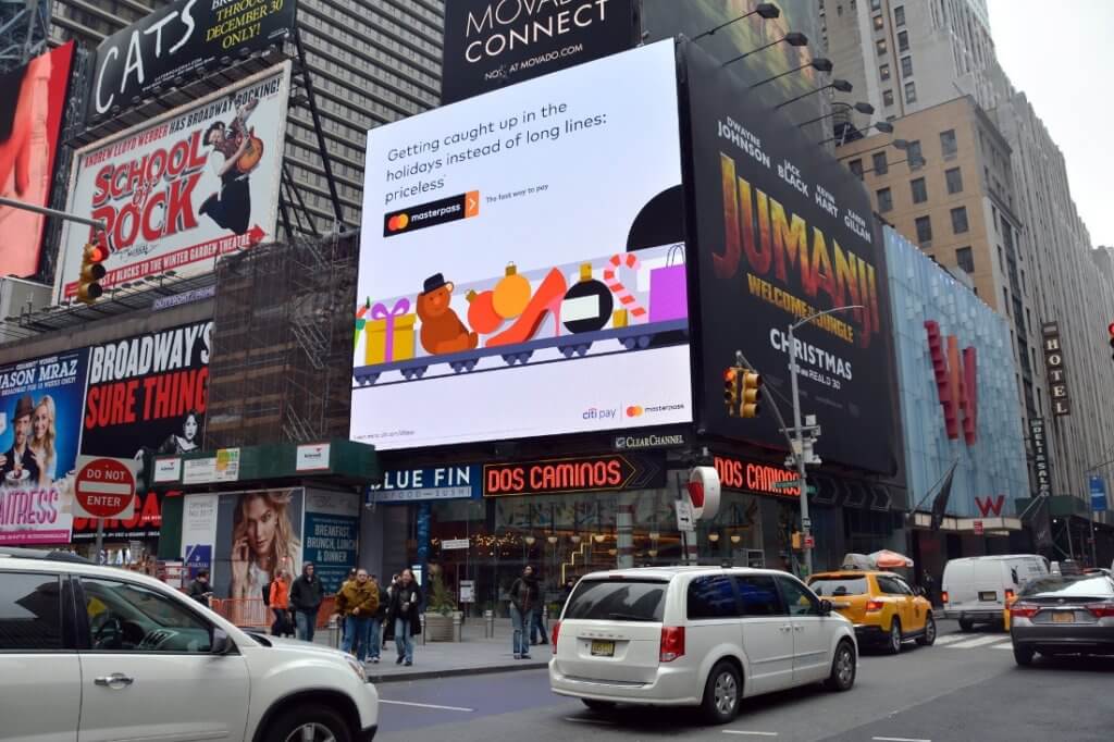 An image of a busy street in downtown New York. The image focuses on a large billboard for Mastercard promoting their holiday shopping online options.