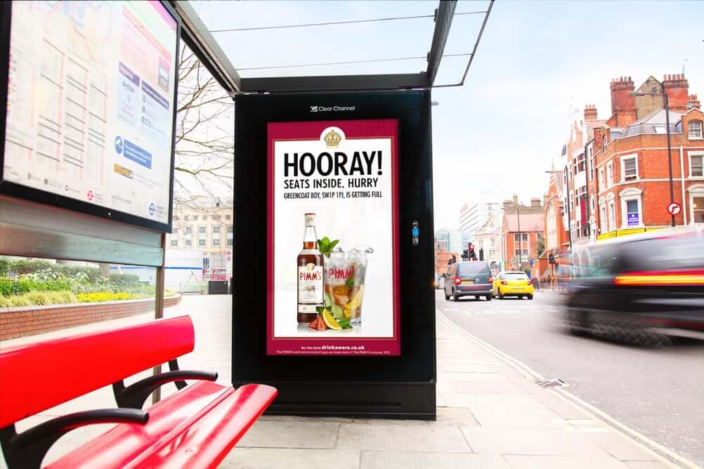 An image of a DOOH ad at a bus stop. The ad is for Pimm's alcohol with a picture of the bottle and the alcohol in a glass with ice and lemons.
