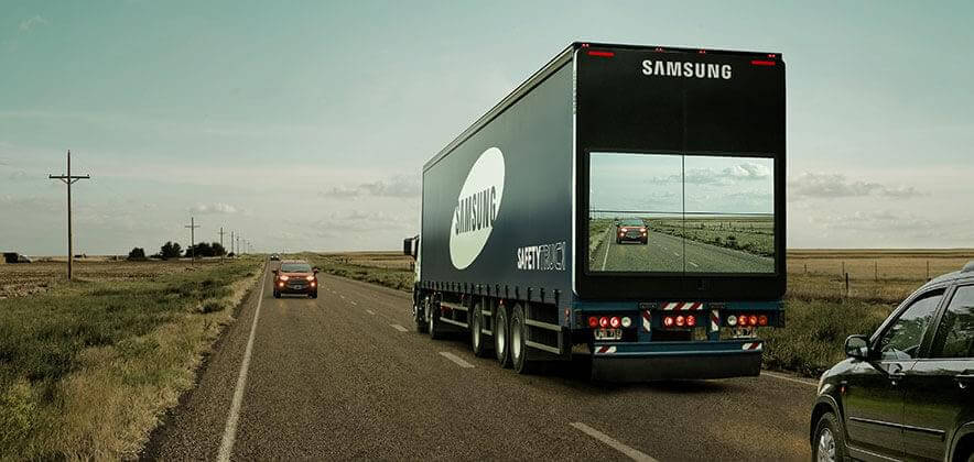 An image of a one-lane highway that focuses on a truck-side ad with a camera on the back of it displaying what is being seen by the truck driver at the front.