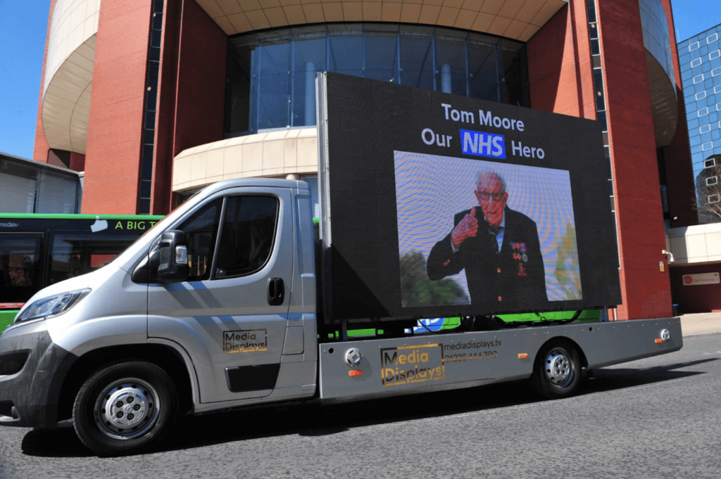 A truck on a road with a huge digital billboard ad on its side showing an image of a veteran and the text reads ' Tom Moore Our NHS Hero'.