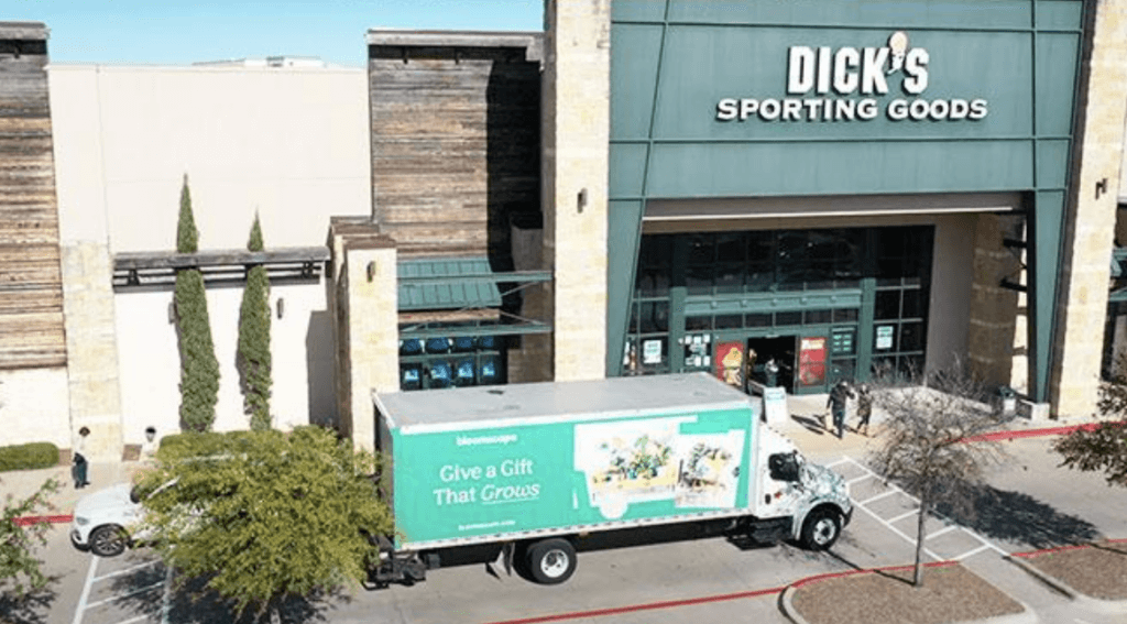 A truck with billboard on the side of the truck that  reads ' Give a Gift That Grows'; parked outside Dick's Sporting Goods complex.