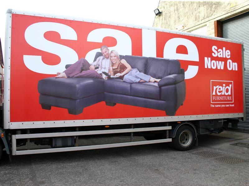 An image of a truck-side ad for a furniture sale at Reid Furniture. Two people laying on a couch laughing.