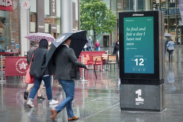 An image showcasing a digital OOH billboard by JCDecaux. The digital billboard displays a Shakespeare quote and the temperature.
