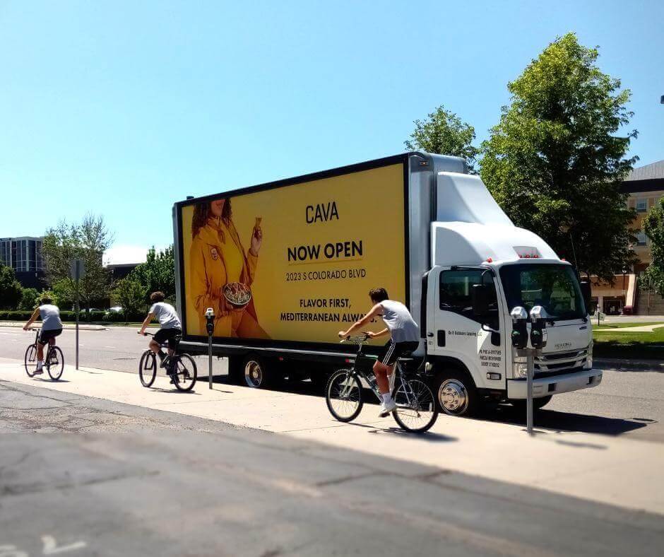 Image of trackside advertisement for CAVA store opening with people biking passing by the truck.