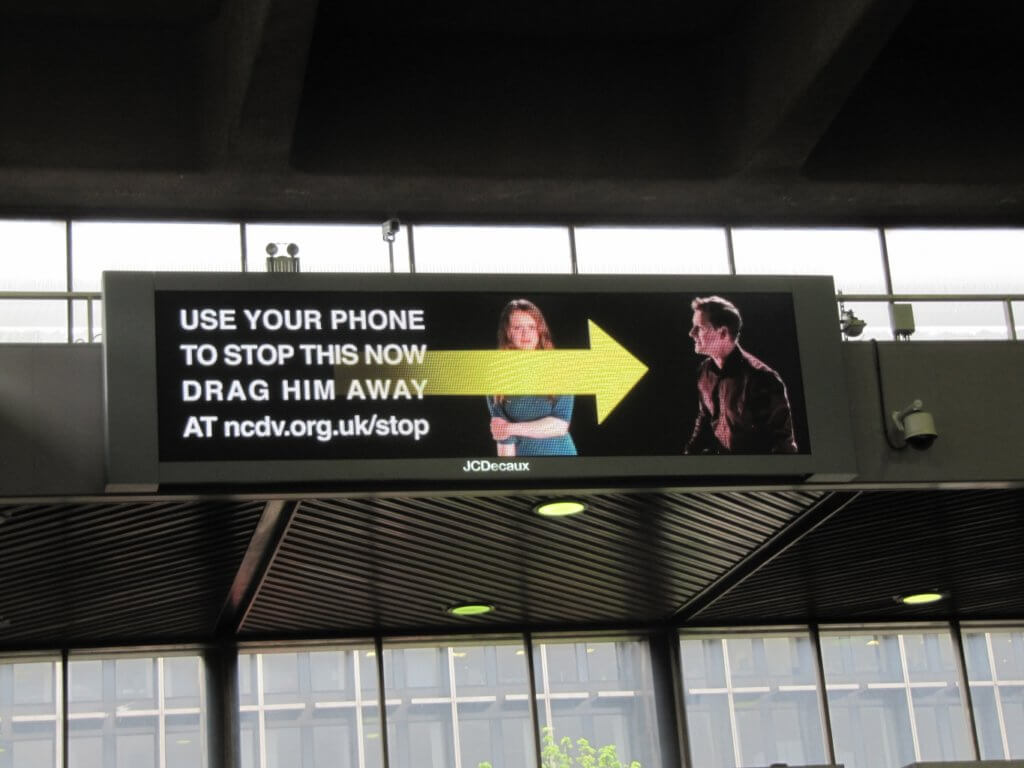 An image of an interactive billboard of a man yelling at a woman with a big yellow arrow encouraging viewers to swipe away the man.