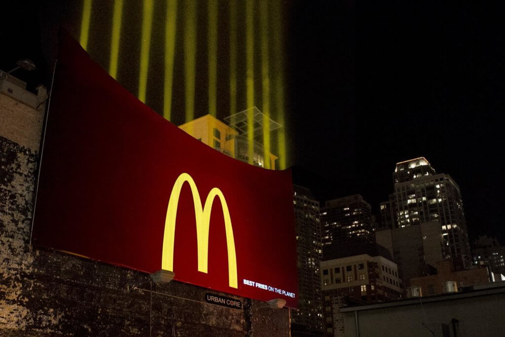 An image of a billboard for McDonald's that just has the golden arches on it but is in the shape of a McDonald's fry container. The billboard has yellow light beams coming out of the top to represent fries.