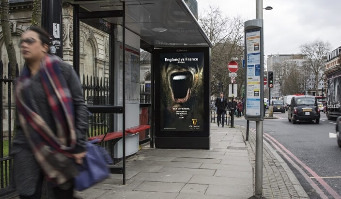 A street corner with a DOOH bus-stop ad for Guinness. The ad displays a mouth thats screaming and tells passersby where they can watch the Six Nations Championship.