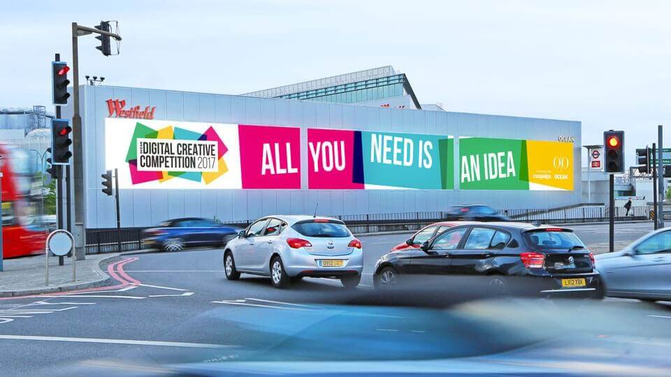 An image of cars in a roundabout passing by a brightly lit and colourful ad that says "All you need is an idea". The ad is for a digital creative competition. 