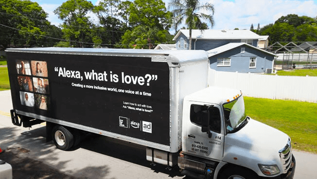 A truck with a mobile billboard on it's side for the product Alexa. The ad shows images of people. The text reads " Alexa, what is love? Creating a more inclusive world, once voice at at time".