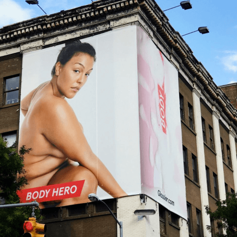 An image of a large-scale ad on the side of a building for Glossier. The ad shows a nude woman showing off her clear skin.