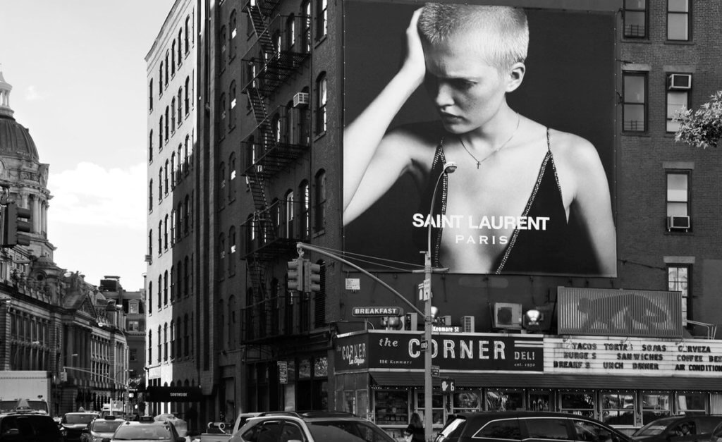 An image of a busy street corner showcasing a Saint Laurent massive billboard. The billboard has a model on it with a low-slung dress, looking down.