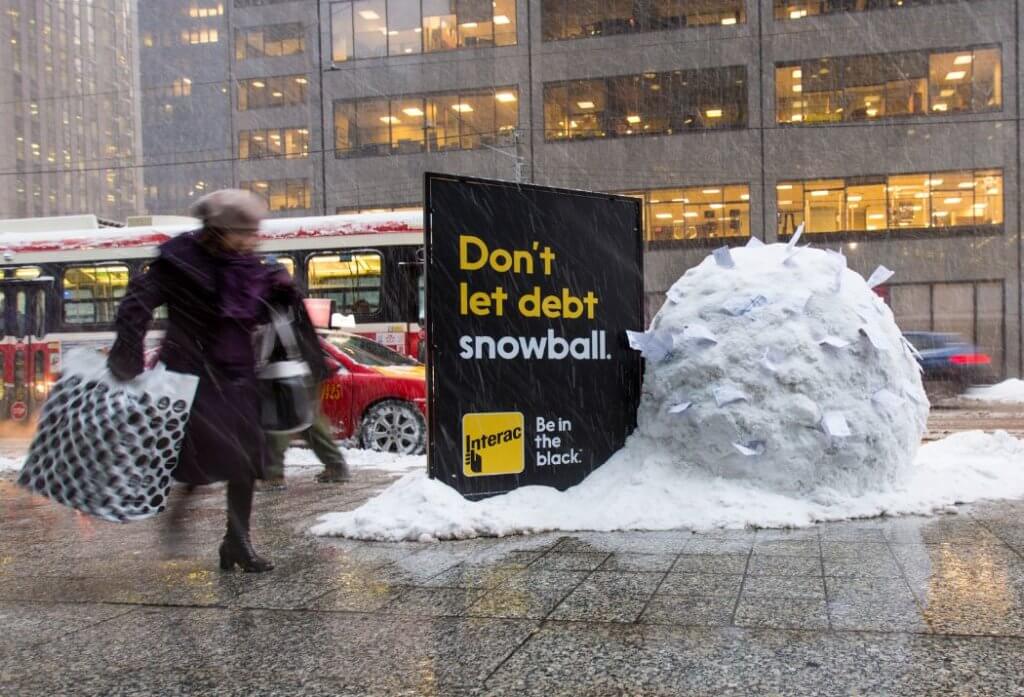 An image of a snowy street that focuses on a billboard for Interac that says, "Don't let debt snowball." with a giant snowball beside it with papers sticking out of it.