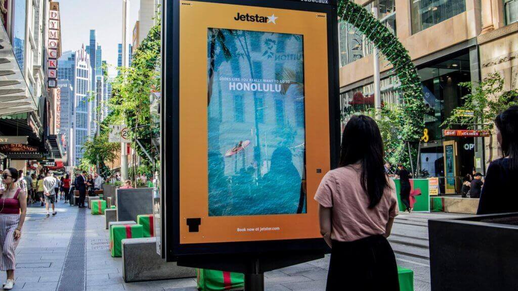 Image of a woman standing and staring at a digital ad by Jetstar that shows someone on a surf board in the middle of the ocean.