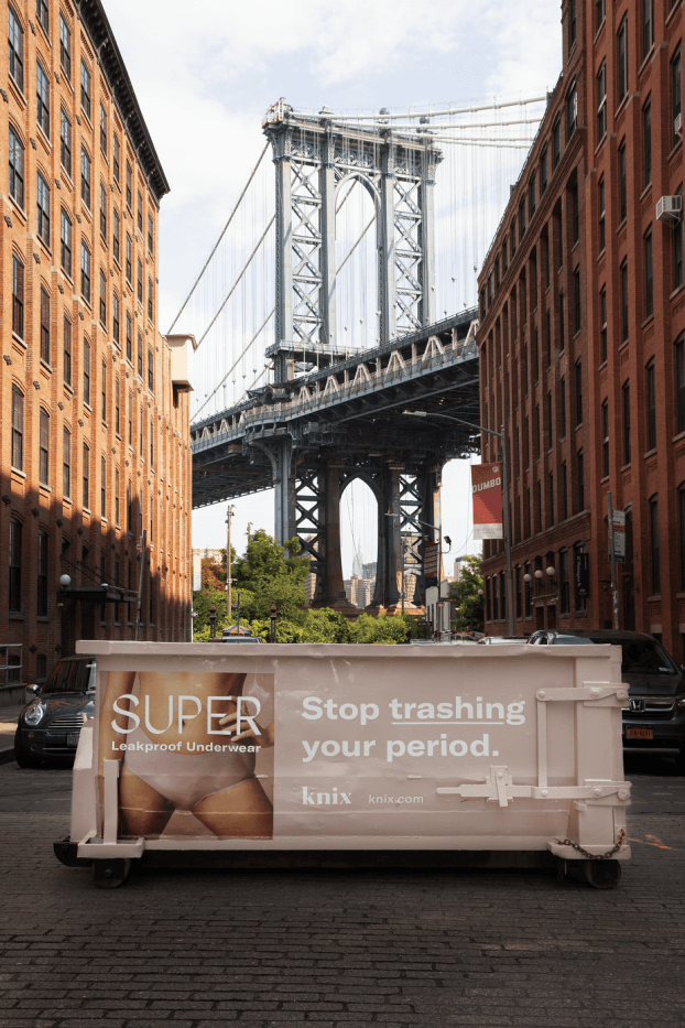 An image of a street in New York City that is showcasing a dumpster truck that has been wrapped with an ad for Knix. On the left hand side it has a woman wearing the new leakproof underwear.