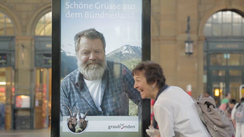 An image of an AOOH advertisement in a train station. The man on the ad is speaking to the woman who is looking at it, she's laughing.