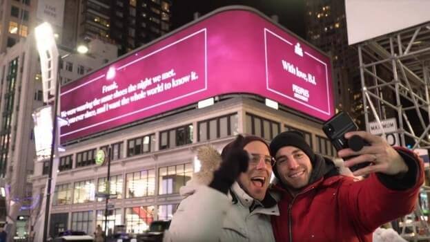 Two men taking a selfie in front of the  Peoples billboard that one of the men wrote for the other.