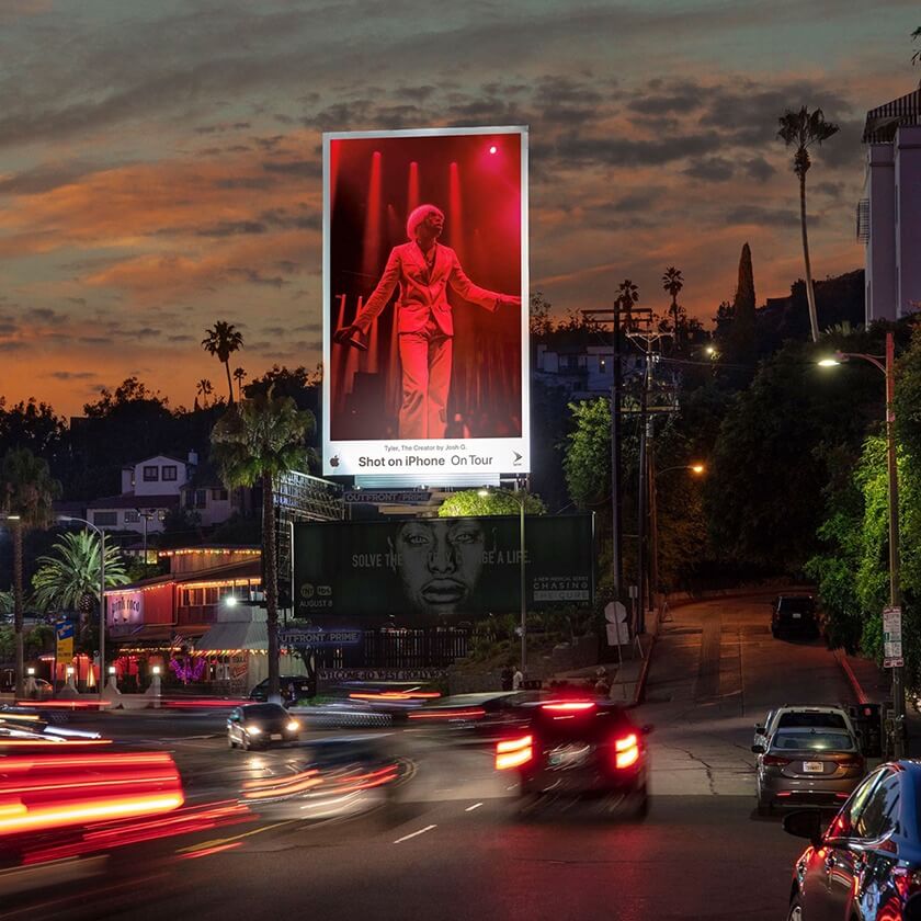 An image of a busy street with a billboard that has an ad for Apple. The ad is a photo of Tyler the Creator in red and he's performing on stage.
