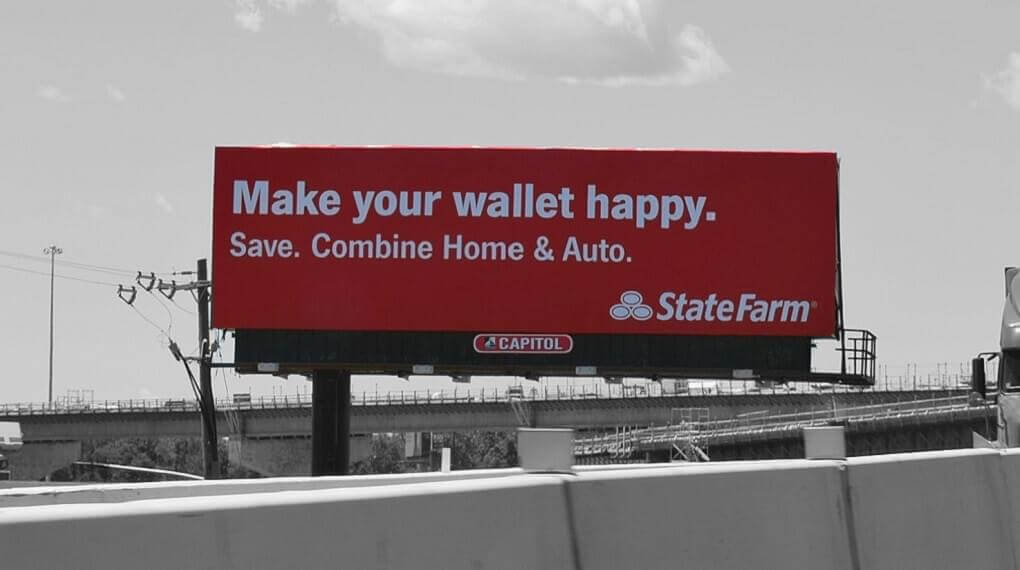 Photo of State Farm billboard advertisement. Advertisement has the brand's signature white slogan against a bright red background. The advertisement says "Make your wallet happy. Save. Combine Home & Auto"
