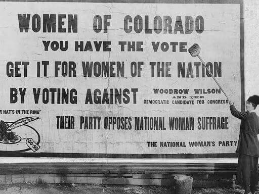 Black and white image of woman gluing up billboard poster in early 1900s for the Suffragette movement. Poster reads, "women of Colorado you have the vote get it for women of the nation by voting against 
Woodrow Wilson.