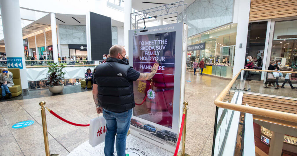 An image of a man in a mall putting his hand up and gesturing to a digital billboard for Skoda.